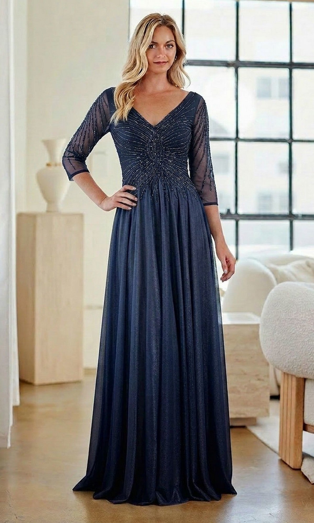A woman stands indoors by a large window, wearing the Amelia Couture KF1501 long formal dress in navy, featuring sheer sleeves, a beaded bodice, and an elegant A-line skirt.